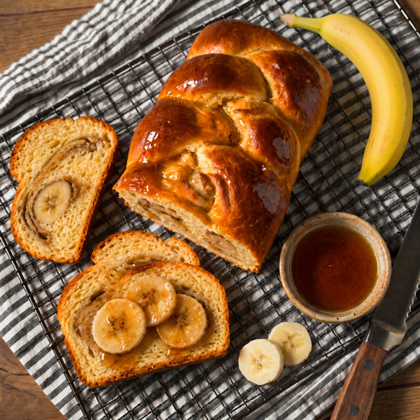 A loaf of glossy banana bread sits on a wire cooling rack on a striped cloth, with two slices topped with banana slices and syrup. A whole banana, a small bowl of syrup, and a serrated knife are arranged around it on a wooden surface.