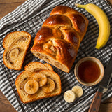 A loaf of glossy banana bread sits on a wire cooling rack on a striped cloth, with two slices topped with banana slices and syrup. A whole banana, a small bowl of syrup, and a serrated knife are arranged around it on a wooden surface.