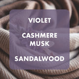 A close-up of soft draped fabric in a muted violet color, overlaid with a semi-transparent purple square containing the text 'VIOLET CASHMERE MUSK SANDALWOOD' in white. The background shows the fabric texture in warm taupe and beige tones.