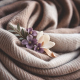 A close-up of soft, draped knit fabric in warm taupe and beige tones, styled with a small floral accent of purple flowers and a cream blossom.