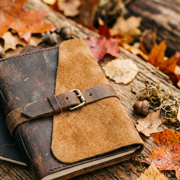 A rustic leather journal with a brown suede flap and metal buckle strap, resting on a weathered wooden surface surrounded by scattered autumn leaves and acorns.