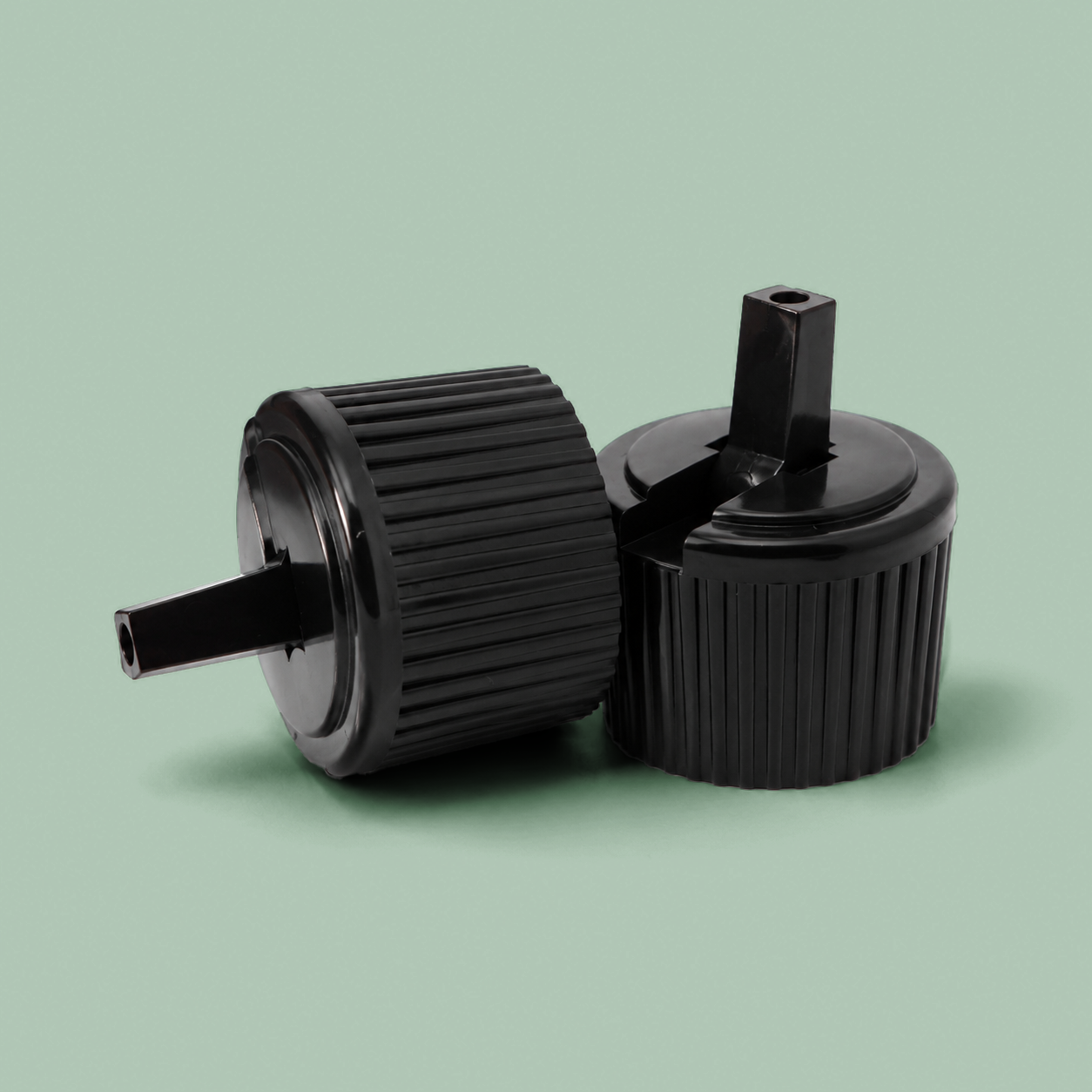Two black plastic bottle caps with ribbed flanges and threaded necks, shown on a solid sage green background.
