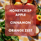 A styled flat-lay of fresh honeycrisp apples, a glass of orange juice, cinnamon sticks, and orange zest on a yellow-and-white checkered cloth, with a translucent red overlay reading 'HONEYCRISP APPLE CINNAMON ORANGE ZEST'.