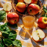 A glass of golden apple juice surrounded by fresh red apples, sliced apples, green leaves, and a wicker basket on a yellow-and-white checkered tablecloth.