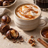A ceramic mug filled with a caramel-topped hot drink sits on a beige tabletop with roasted chestnuts, a cinnamon stick, coarse salt, and a small dish of caramel. 
