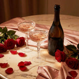 Two glasses of pink champagne, a bottle, and red roses on a marble surface with a pink cloth.