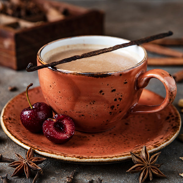 A warm terracotta ceramic cup filled with a light brown beverage, set on a matching saucer with two fresh cherries, star anise, cinnamon sticks, and a vanilla bean in a rustic styled setting.