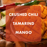 A close-up food photo of diced orange-red mango with visible crushed chili powder, overlaid with a translucent orange label reading 'CRUSHED CHILI TAMARIND MANGO.' Surrounding ingredients include lime wedges, a red chili, green chili, and other fruits on a wooden surface.