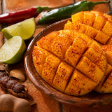 Cubed mango slices seasoned with red chili powder, served in a brown bowl with lime wedges, whole chilies, and tamarind on a rustic wooden surface.