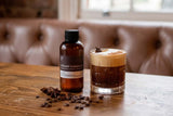 Bottle of 'Creamy Carajillo' Fragrance2U fragrance oil with a glass of layered coffee drink on a wooden table.