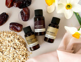 Three small fragrance oil bottles with labels next to dried oats, dates, and flowers on a light surface.