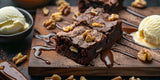 Two walnut brownies lay on a wooden plate surrounded by chocolate drizzle, cocoa powder, and ice cream.