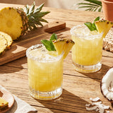 Two glasses of pineapple cocktail on a rustic wooden table, garnished with fresh pineapple wedges and mint leaves, with sliced pineapples and coconut shells nearby.
