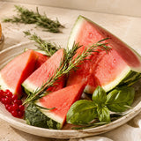 Slices of red watermelon arranged in a shallow bowl with green rinds, garnished with fresh rosemary sprigs and basil leaves, on a light beige fabric background.