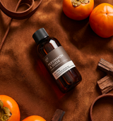 Bottle of 'Pomegranate & Suede' Fragrance2U fragrance oil on a brown textured surface with persimmons and wood pieces.