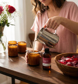 Person pouring wax into a candle on a wooden table with flowers and a bottle of rosewater & Oud Fragrance2U fragrance oil in the background