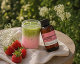 Candle with green and pink gradient, strawberry milk & matcha fragrance oil bottle, and strawberries on a wooden surface with a natural background.