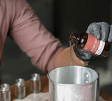Person pouring liquid from a bottle labeled 'Sultry Santal' into a metal container for making scented products