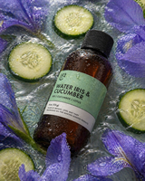 Bottle of 'Water Iris & Cucumber' fragrance oil surrounded by sliced cucumbers and purple flowers on a reflective surface.